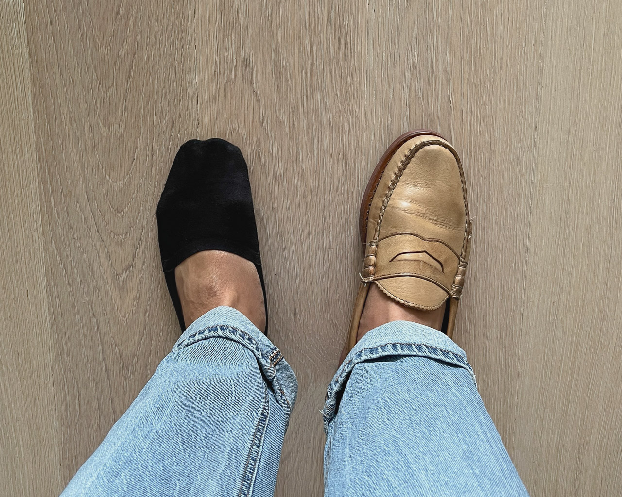 top-down closeup shot of man wearing light blue jeans and one tan loafer on the right foot with one ondo black no show sock on the left foot