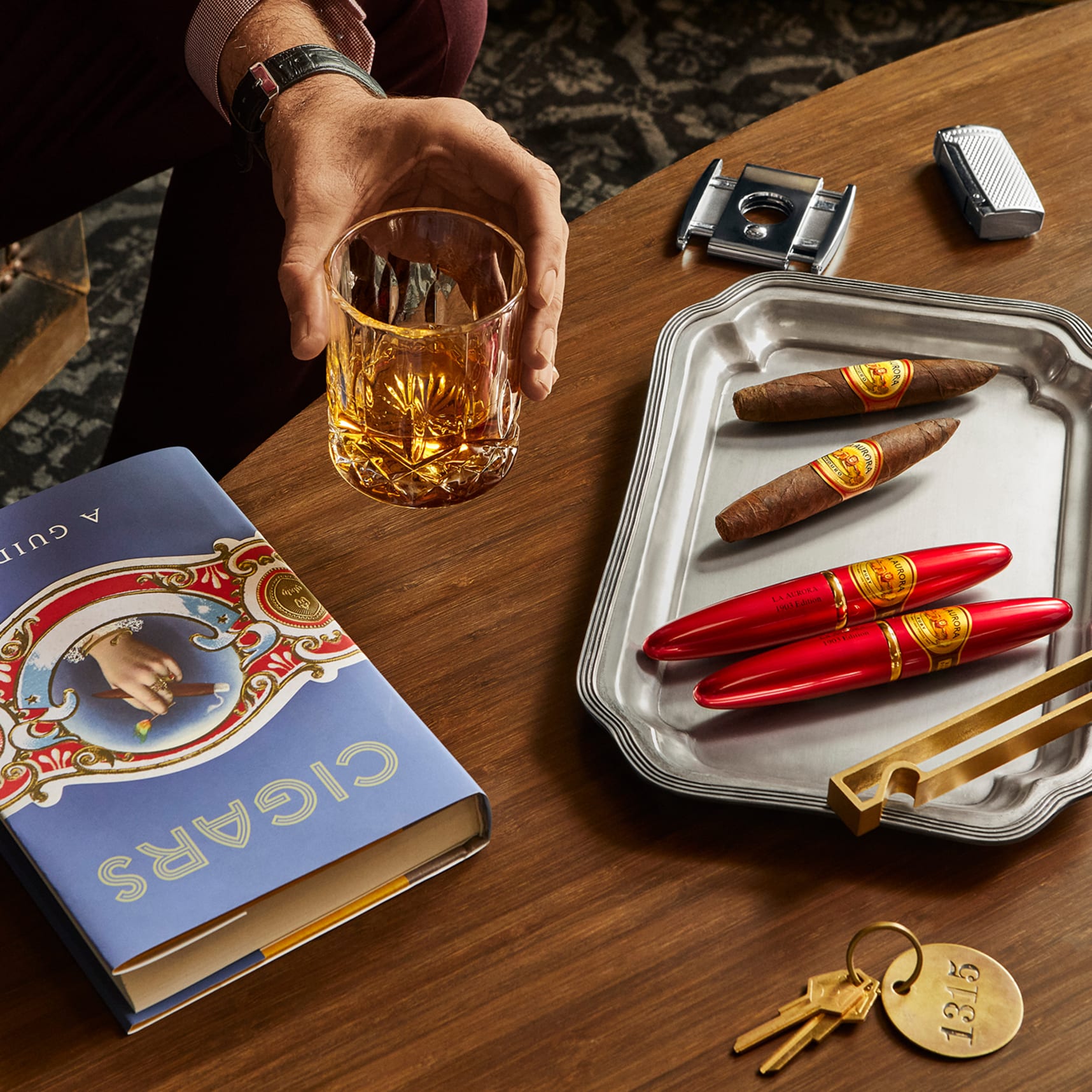 close up of tray with red cigar tubes and man's hand holding bourbon glass next to cigar book